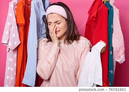Crying woman keeps right hand near her face, holds hanger with white shirt while standing at her wardrobe. Dark haired woman has nothing to wear and no money for buying new outfit. People, problems. 76592801