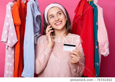Young woman with credit card dreams about shopping while standing near lots hangers with colourful clothes isolated over pink background. Fashoin male likes online shopping. Smiling girl buys dress. Young woman with credit card dreams about shopping while standing near lots hangers with colourful clothes isolated over pink background. Fashoin male likes online shopping. Smiling girl buys dress. 76592802