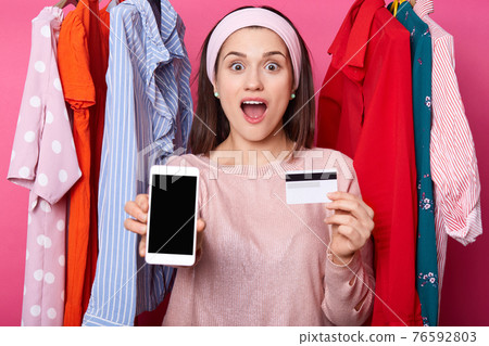 Portrait of excited young dark haired girl shows plastic credit card which full of money while holding mobile phone isolated over rose background. Smiling female shows phone, woman gets salary. Portrait of excited young dark haired girl shows plastic credit card which full of money while holding mobile phone isolated over rose background. Smiling female shows phone, woman gets salary. 76592803
