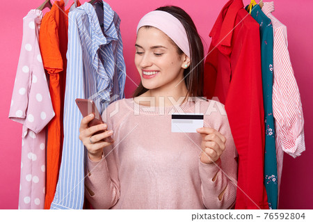 Beautiful dark haired young female with cheerful expression, holds smart phone and credit card. Happy girl makes online payment. Woman chooses outfits in online shop. Payment and shopping concept. Beautiful dark haired young female with cheerful expression, holds smart phone and credit card. Happy girl makes online payment. Woman chooses outfits in online shop. Payment and shopping concept. 76592804