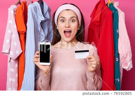 Photo of screaming young lady in clothes store. Girl with opened mouth holds mobile phone and credit card. Female near hagers with colourful outfits. Woman gets message with ballance on card. 76592805