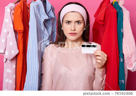 Dicontent woman holds credit card and looks stressed, she has financial problems. Brunette female spends all money from her card. Model poses against hangers with colourfuul dresses. Shopping concept. Dicontent woman holds credit card and looks stressed, she has financial problems. Brunette female spends all money from her card. Model poses against hangers with colourfuul dresses. Shopping concept. 76592806