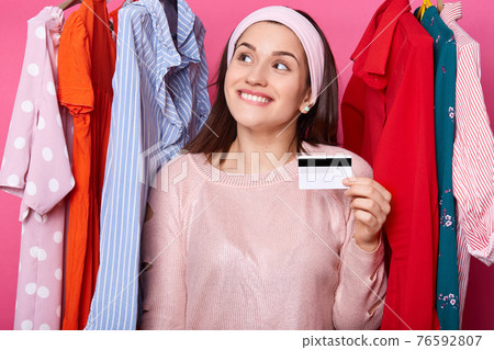 Happy female holds a credit card and smiles. Young shopper feels goos, bites her lip. Attractive woman has unlimited plastic card for buying dresses in big shopping mall. Shopping and fashion concept. 76592807