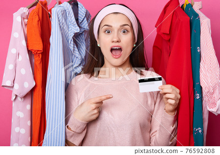 Suprised woman with opened mouth holds credit card. Lady wears pink sweater points aside at card. Glad female has money for shopping. Young girl chooses clothes in mall. Fasion, shopping concept. 76592808