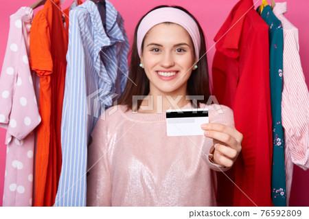 Smiling yuong dark haired woman holds credit card. Lady has unlimited card for buying dresses. Happy female can pay for purchase by card. Cheerful girl near hangers with outfits. Shopping concept. Smiling yuong dark haired woman holds credit card. Lady has unlimited card for buying dresses. Happy female can pay for purchase by card. Cheerful girl near hangers with outfits. Shopping concept. 76592809