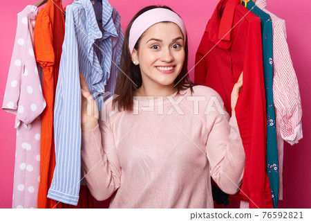 Happy smiling girl stands between hangers with blouses in fashion store. Cheerful woman visits boutique. Cute lady likes to go to mall. Beautiful young female on sale. Shopping and fasion concept. 76592821