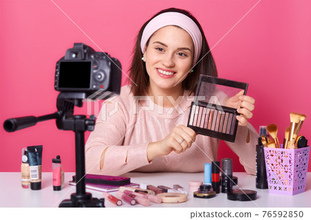 Portrait of young successful trendy blogger sitting in front of camera, showing palette of eyeshadow, holding it in hand, wearing headband and fashionable pink sweater, looks delighted. Shooting. 76592850