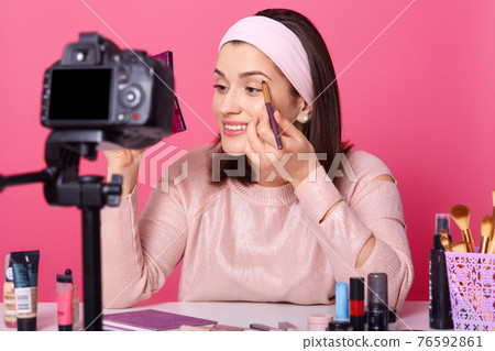 Beautiful female records video. Yong blogger shows how to apply eyeshadow. Lady advetises cosmetics on her channel. Woman holds mirror and brush against pink wall. Fasion and blogging concept. Beautiful female records video. Yong blogger shows how to apply eyeshadow. Lady advetises cosmetics on her channel. Woman holds mirror and brush against pink wall. Fasion and blogging concept. 76592861