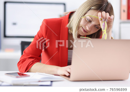 Tired woman sitting at laptop with glasses in her hands 76596899