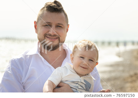 Happy father on vacation - smiling man standing on a beach holding his little baby son Happy father on vacation - smiling man standing on a beach holding his little baby son 76597150