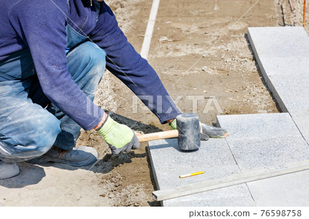A worker places granite blocks on the sidewalk with a rubber hammer on a marked area. 76598758