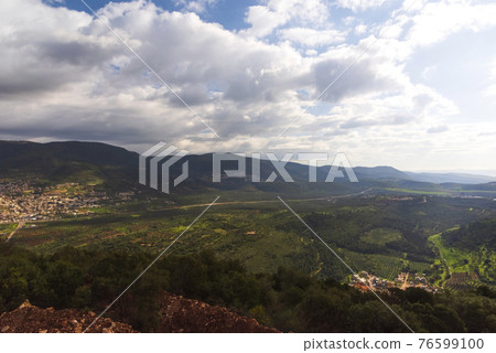 A view of a mountain range and a green valley in the morning at sunrise, against a dramatic back of blue skies and clouds. High quality photo. Travel concept hiking. North District Israel A view of a mountain range and a green valley in the morning at sunrise, against a dramatic back of blue skies and clouds. High quality photo. Travel concept hiking. North District Israel 76599100