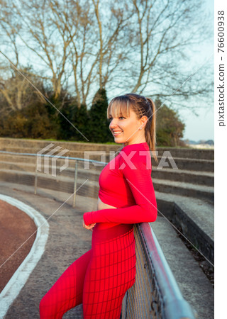 Portrait of a smiling confident fitness woman in red sport wear resting after work out or running outdoors on sunset. Jogging in the stadium. Active lifestyle and health care. Vertical card. Portrait of a smiling confident fitness woman in red sport wear resting after work out or running outdoors on sunset. Jogging in the stadium. Active lifestyle and health care. Vertical card. 76600938