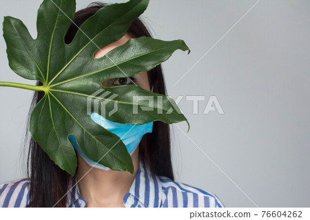 Beautiful woman hides face in a medical mask and open her eyes with big green tropical leaf, Faceless, Protection from covid 76604262