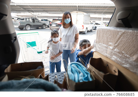 Asian mother and two daughters wearing protection mask helping to put stuffs in the shopping trolley into trunk of car together in parking during coronavirus pandemic as new normal lifestyle. 76608685
