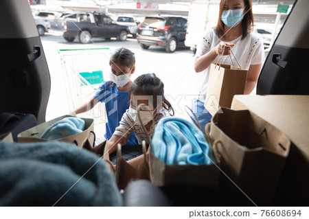 Asian mother and two daughters wearing protection mask helping to put stuffs in the shopping trolley into trunk of car together in parking during coronavirus pandemic as new normal lifestyle. Asian mother and two daughters wearing protection mask helping to put stuffs in the shopping trolley into trunk of car together in parking during coronavirus pandemic as new normal lifestyle. 76608694