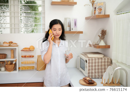 Portrait of smiling young beautiful woman standing, drinking water and talking on smartphone in kitchen. 76615174