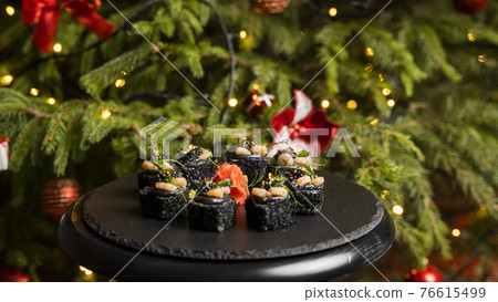 Sushi with black rice, crab meat, avocado, smoked salmon mousse, oar caviar, masago, shrimp cocktail and edible gold leaf with ginger on black table for Christmas with a Christmas tree on background. 76615499