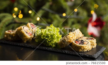 Custom sushi roll in tempura with nori, fresh salmon, tuna, avocado, masago caviar, drizzled with pineapple sauce with salad pouring as decoration on a black plate on a wooden table for Christmas. 76615500