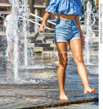A young girl walks barefoot along the granite surface of the city fountain among the pouring water on a bright hot day. A young girl walks barefoot along the granite surface of the city fountain among the pouring water on a bright hot day. 76627320