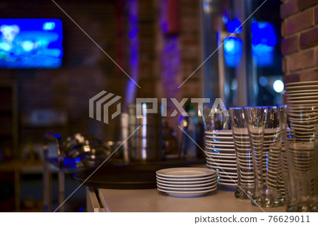 Stacks of a clean shiny utensils made of steel, ceramics and glass in a room with blue of light in the TV and window. Stacks of a clean shiny utensils made of steel, ceramics and glass in a room with blue of light in the TV and window. 76629011