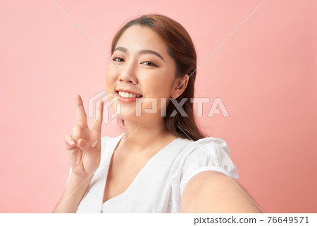 Happy woman with beaming smile making selfie and showing v-sign against pink background 76649571