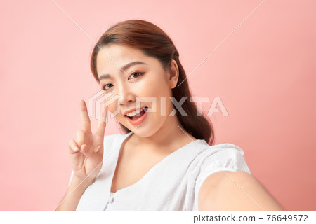 Happy expressive young woman posing while taking pictures of herself through cellphone, over pink background 76649572