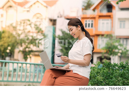 Photo of happy asian woman using laptop and holding credit card while sitting on bench in green park 76649623