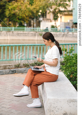 Casual girl buying online with a laptop and paying with a credit card sitting on a bench outdoors in the street 76649629