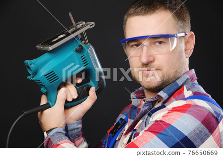 Caucasian man builder carpenter in a shirt and overalls with an electric tool in his hands. Studio portrait of friendly artisan businessman 76650669