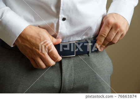A man in blue pants and a white shirt buttoned a brown leather trouser belt. He has a watch on his hand. 76663874