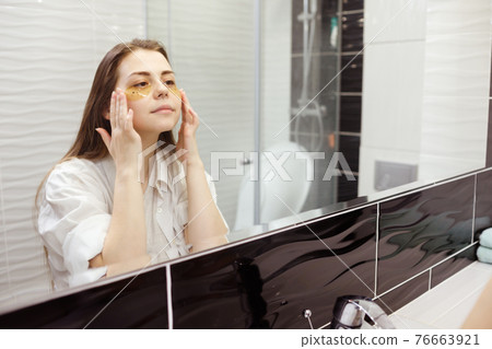 Caucasian woman applying hydrogel patches on under eye area, looking at mirror. Happy young lady enjoying morning anti age anti wrinkle routine in bathroom Caucasian woman applying hydrogel patches on under eye area, looking at mirror. Happy young lady enjoying morning anti age anti wrinkle routine in bathroom 76663921