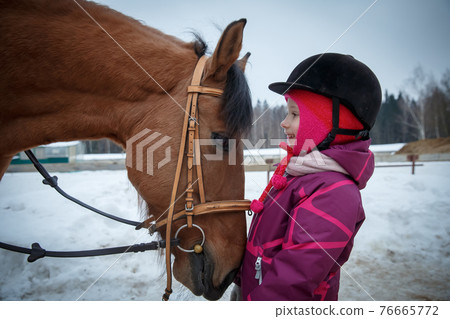 Close up of a little girl jockey smiling and looking into the horse eyes, in the profile. Girl horse rider with a horse in a winter open riding hall, looking eye to eye. Girl and horse friendship, equ 76665772