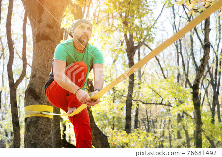 A bearded man in age balances while sitting on a taut slackline in the autumn forest. Outdoor Leisure A bearded man in age balances while sitting on a taut slackline in the autumn forest. Outdoor Leisure 76681492