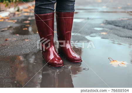 Female feet in red rubber boots stand in a muddy puddle under rain in autumn weather Female feet in red rubber boots stand in a muddy puddle under rain in autumn weather 76688504