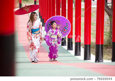 woman and little girl in kimono holding umbrella walking into at the shrine red gate, in Japanese garden. 76689044