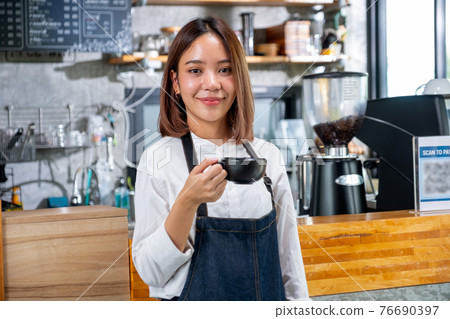 Portrait of barista woman or coffee maker hold a cup and stand in front of counter in coffee shop Portrait of barista woman or coffee maker hold a cup and stand in front of counter in coffee shop 76690397