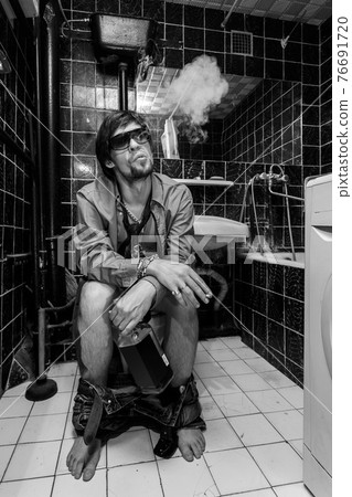 Drunk Man sits in a toilet with a bottle of whiskey and cigarette Drunk Man sits in a toilet with a bottle of whiskey and cigarette 76691720
