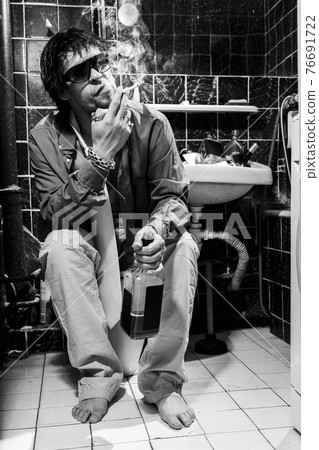 Drunk Man sits in a toilet with a bottle of whiskey and cigarette Drunk Man sits in a toilet with a bottle of whiskey and cigarette 76691722