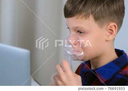 Boy is breathing in mask using nebulizer for inhalation at home watching laptop. Boy is breathing in mask using nebulizer for inhalation at home watching laptop. 76691933