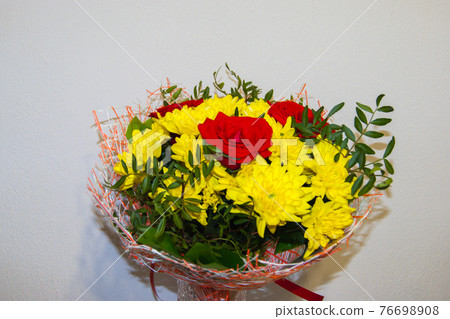 A bouquet of yellow chrysanthemums and red roses in the package stands in a vase against the white A bouquet of yellow chrysanthemums and red roses in the package stands in a vase against the white 76698908