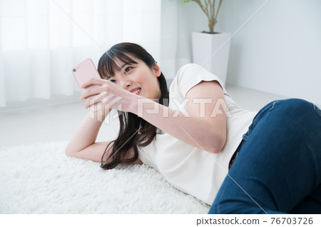 A woman lying on a carpet and looking at her smartphone 76703726