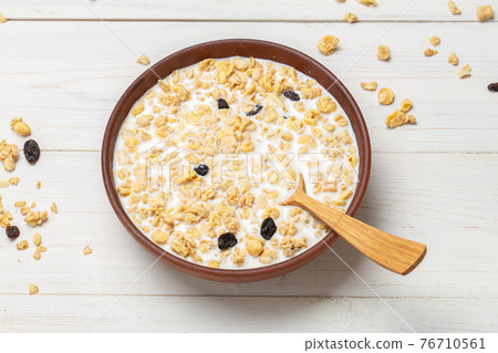 Muesli with dried grapes is poured with milk in a clay bowl on a white wooden table. 76710561