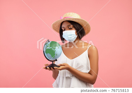 Travel ban during covid lockdown. Sad black woman in summer dress wearing face mask and holding globe on pink background 76715813