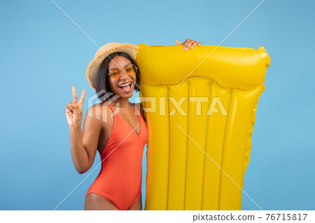 Cheerful black lady in swimsuit holding yellow lilo and showing peace gesture on blue studio background Cheerful black lady in swimsuit holding yellow lilo and showing peace gesture on blue studio background 76715817