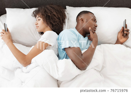Couple using phones in bed, lying back to back, ignoring after quarrel Couple using phones in bed, lying back to back, ignoring after quarrel 76715976