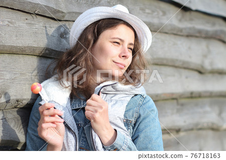 Portrait of dreamy girl with a lollipop near wooden fence. Beautiful young caucasian woman with sweet Chupa-Chups. Spring holidays in ferme. 76718163