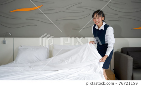 Young hotel maid with a smile making the bed. Staff in blue uniform preparing room for hotel guest. Young hotel maid with a smile making the bed. Staff in blue uniform preparing room for hotel guest. 76721159