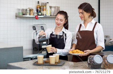 Young asian barista with a smile pouring milk from the jug into a paper coffee cup. Caucasian assistant in an apron holding croissant plate stands beside her. Morning atmosphere in a coffee shop. Young asian barista with a smile pouring milk from the jug into a paper coffee cup. Caucasian assistant in an apron holding croissant plate stands beside her. Morning atmosphere in a coffee shop. 76721279