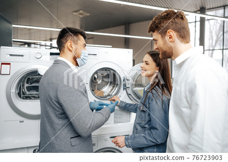 Salesman in hypermarket wearing medical mask demonstrates his clients a new washing machine Salesman in hypermarket wearing medical mask demonstrates his clients a new washing machine 76723903